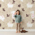 Lovely child standing in front of Whimsy Tots Bunny and Butterfly Meadow Wallpaper in Beige with rabbit, butterfly, bunny, branch, leaf in design.