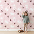 Lovely child standing in front of Whimsy Tots Bunny and Butterfly Meadow Wallpaper in Blush Pink with rabbit, butterfly, bunny, branch, leaf in design.