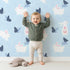 Lovely child standing in front of Whimsy Tots Bunny and Butterfly Meadow Wallpaper in Blue with rabbit, butterfly, bunny, branch, leaf in design.