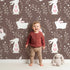 Lovely child standing in front of Whimsy Tots Tribal Bunny Sprigs Wallpaper in Cocoa Brown with rabbit, bunny, sprig, leaf, tribal motif in design.