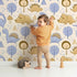 Lovely child standing in front of Whimsy Tots Jurassic Forest Friends Wallpaper in Cream with dinosaur, tree, triangle, dino, forest in design.