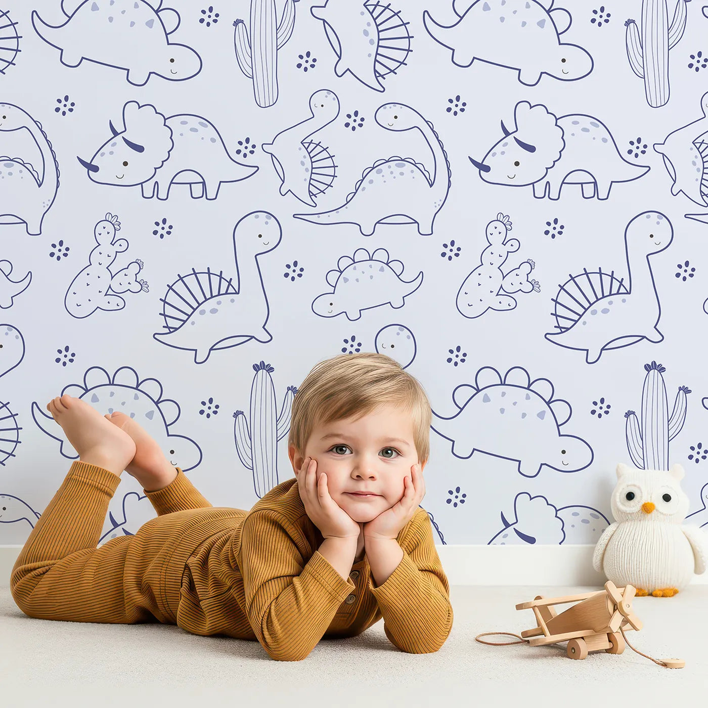 Lovely child standing in front of Whimsy Tots Desert Dino Parade Wallpaper in Sky Blue with dinosaur, cactus, flower in design.