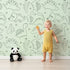 Lovely child standing in front of Whimsy Tots Desert Dino Parade Wallpaper in Sage Green with dinosaur, cactus, flower in design.