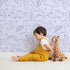 Lovely child standing in front of Whimsy Tots Desert Dino Parade Wallpaper in Lavender with dinosaur, cactus, flower in design.