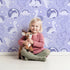 Lovely child standing in front of Whimsy Tots Dino Circle Parade Wallpaper in Lavender with dinosaur, flower, heart, circle in design.