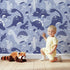 Lovely child standing in front of Whimsy Tots Botanic Dino Meadow Wallpaper in Periwinkle with dinosaur, flower, cactus, leaf in design.