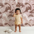 Lovely child standing in front of Whimsy Tots Botanic Dino Meadow Wallpaper in Rosewood Pink with dinosaur, flower, cactus, leaf in design.