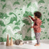 Lovely child standing in front of Whimsy Tots Botanic Dino Meadow Wallpaper in Sage Green with dinosaur, flower, cactus, leaf in design.