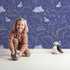 Lovely child standing in front of Whimsy Tots Midnight Dino Parade Wallpaper in Navy Blue with dinosaur, cactus, heart, footprint in design.
