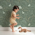 Lovely child standing in front of Whimsy Tots Midnight Dino Parade Wallpaper in Forest Green with dinosaur, cactus, heart, footprint in design.