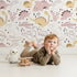 Lovely child standing in front of Whimsy Tots Cactus Canyon Dinos Wallpaper in Blush Pink with dinosaur, cactus, rock, spike in design.
