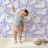 Lovely child standing in front of Whimsy Tots Swan Princess Waltz Wallpaper in Lavender with swan, crown, wing, heart, star in design.