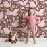Lovely child standing in front of Whimsy Tots Starlit Swan Ballet Wallpaper in Cocoa Brown with swan, crown, flower, bow, leaf in design.