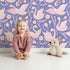 Lovely child standing in front of Whimsy Tots Starlit Swan Ballet Wallpaper in Periwinkle Purple with swan, crown, flower, bow, leaf in design.