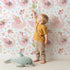 Lovely child standing in front of Whimsy Tots Blooming Swan Garden Wallpaper in White with swan, flower, crown, tulip, ripple in design.