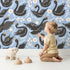 Lovely child standing in front of Whimsy Tots Regal Swan Parade Wallpaper in Sky Blue with swan, flower, crown, tulip, leaf in design.