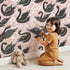 Lovely child standing in front of Whimsy Tots Regal Swan Parade Wallpaper in Blush Pink with swan, flower, crown, leaf in design.