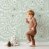 Lovely child standing in front of Whimsy Tots Spring Meadow Whispers Wallpaper in Sage Green with flower, vine, leaf, dot in design.