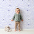 Lovely child standing in front of Whimsy Tots Spring Meadow Whispers Wallpaper in Lavender with flower, vine, leaf, dot in design.