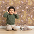 Lovely child standing in front of Whimsy Tots Floral Snuggle Bears Wallpaper in Mocha Brown with bear, flower, heart, leaf in design.