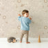 Lovely child standing in front of Whimsy Tots Floral Fox Cuddles Wallpaper in Taupe Beige with fox, flower, leaf in design.