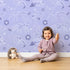 Lovely child standing in front of Whimsy Tots Floral Fox Cuddles Wallpaper in Lavender Mist with fox, flower, leaf in design.