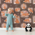 Lovely child standing in front of Whimsy Tots Fox Meadow Moments Wallpaper in Cocoa Brown with fox, flower, frame in design.