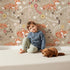 Lovely child standing in front of Whimsy Tots Folk Bloom Foxes Wallpaper in Sandstone Beige with fox, flower, vine in design.