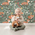 Lovely child standing in front of Whimsy Tots Folk Bloom Foxes Wallpaper in Forest Green with fox, flower, vine in design.