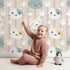 Lovely child standing in front of Whimsy Tots Pawprint Kitty Parade Wallpaper in Oatmilk Beige with cat, pawprint, stripe in design.
