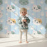 Lovely child standing in front of Whimsy Tots Pawprint Kitty Parade Wallpaper in Baby Blue with cat, pawprint, stripe in design.