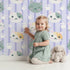 Lovely child standing in front of Whimsy Tots Pawprint Kitty Parade Wallpaper in Lavender Mist with cat, pawprint, stripe in design.