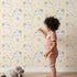 Lovely child standing in front of Whimsy Tots Cozy Pawstep Kittens Wallpaper in Beige with cat, kitten, pawprint, spot in design.