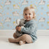 Lovely child standing in front of Whimsy Tots Cozy Pawstep Kittens Wallpaper in Sky Blue with cat, kitten, pawprint, spot in design.