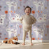 Lovely child standing in front of Whimsy Tots Floral Kitty Parade Wallpaper in Lavender Mist with cat, flower, branch, petal in design.