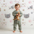 Lovely child standing in front of Whimsy Tots Whisker Wink Kitty Wallpaper in White with cat, pawprint, whisker, smile in design.