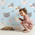 Lovely child standing in front of Whimsy Tots Whisker Wink Kitty Wallpaper in Sky Blue with cat, pawprint, whisker, smile in design.