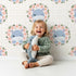 Lovely child standing in front of Whimsy Tots Blossom Hippo Winks Wallpaper in Buttercream Plaid with hippo, flower, wreath, rose in design.