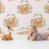 Lovely child standing in front of Whimsy Tots Floral Bear Hugs Wallpaper in Blush Pink with bear, flower, crown, branch, leaf in design.
