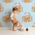 Lovely child standing in front of Whimsy Tots Floral Bear Hugs Wallpaper in Sky Blue with bear, flower, crown, branch, leaf in design.