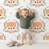Lovely child standing in front of Whimsy Tots Floral Fox Peek Wallpaper in Soft Peach with fox, flower, crown, branch, leaf, polka in design.