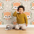 Lovely child standing in front of Whimsy Tots Floral Fox Peek Wallpaper in Mushroom Taupe Beige with fox, flower, crown, leaf, polka in design.