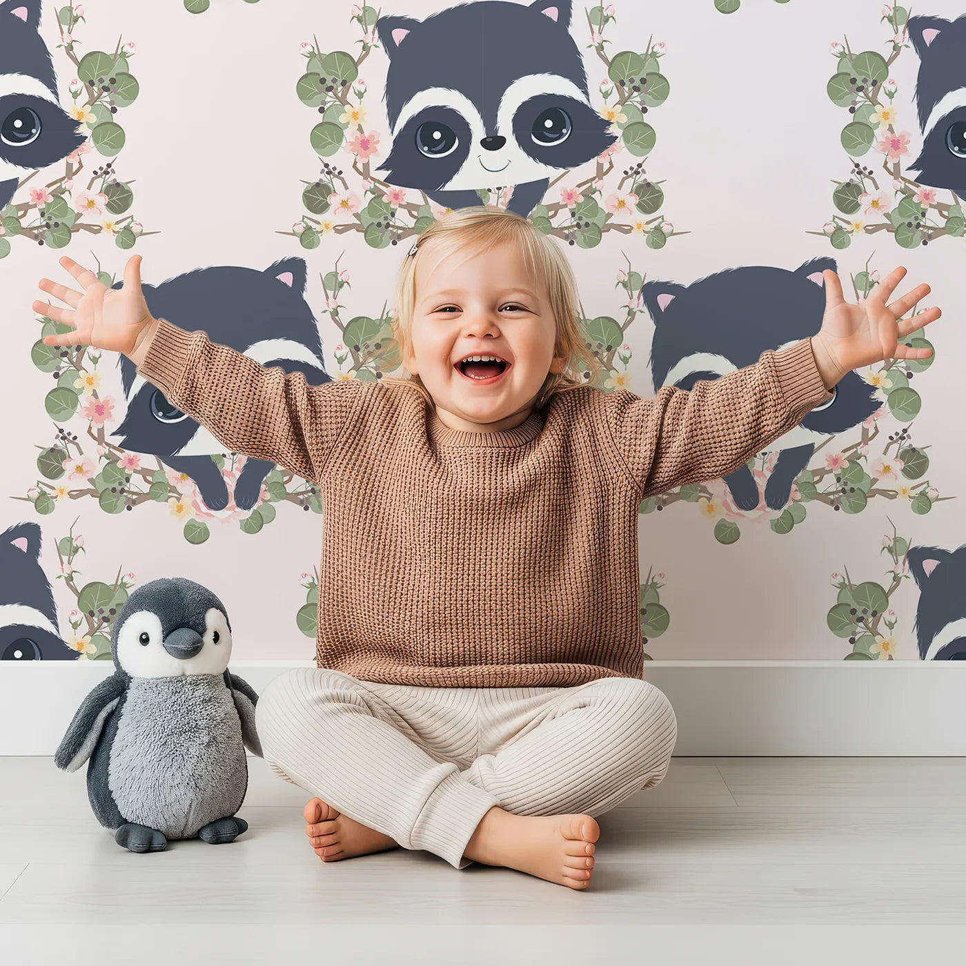 Lovely child standing in front of Whimsy Tots Floral Raccoon Peek Wallpaper in Soft Blush Pink with raccoon, flower, crown, branch, leaf in design.