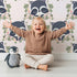Lovely child standing in front of Whimsy Tots Floral Raccoon Peek Wallpaper in Soft Blush Pink with raccoon, flower, crown, branch, leaf in design.