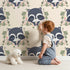 Lovely child standing in front of Whimsy Tots Floral Raccoon Peek Wallpaper in Soft Cream with raccoon, flower, crown, leaf, branch in design.