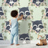 Lovely child standing in front of Whimsy Tots Floral Raccoon Peek Wallpaper in Mint Green with raccoon, flower, crown, leaf, branch in design.