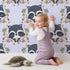 Lovely child standing in front of Whimsy Tots Floral Raccoon Peek Wallpaper in Lavender Mist with raccoon, flower, crown, branch, leaf in design.