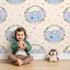 Lovely child standing in front of Whimsy Tots Floral Elephant Wink Wallpaper in Vanilla Cream with elephant, heart, flower, crown, branch, leaf in design.