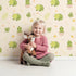 Lovely child standing in front of Whimsy Tots Hatching Dino Friends Wallpaper in Warm Cream with dinosaur, egg, leaf, flower, bone in design.