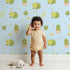 Lovely child standing in front of Whimsy Tots Hatching Dino Friends Wallpaper in Sky Blue with dinosaur, egg, leaf, flower, bone in design.